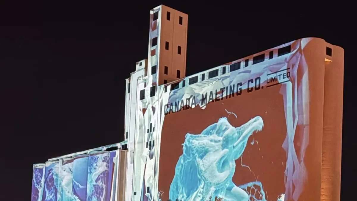Dissolving Boundaries on the night: a pale blue liquid figure projected across the Canada Malting Co. facade, with water imagery running down the silo bins